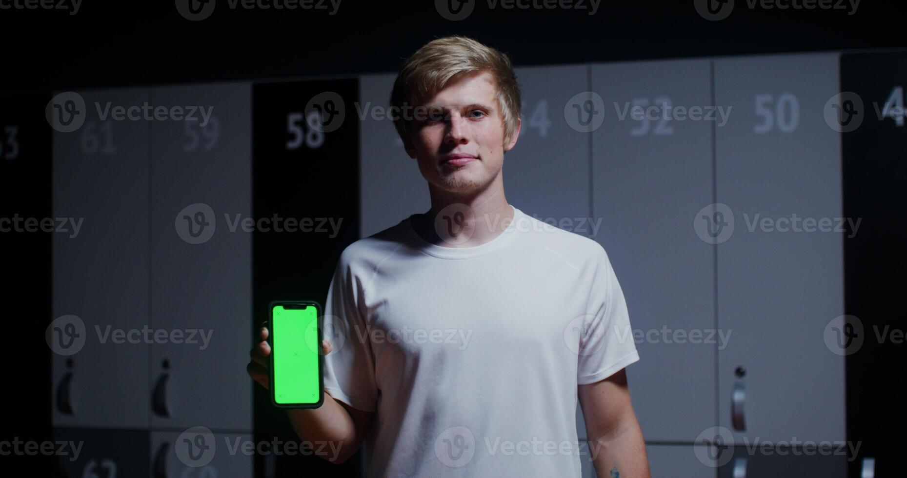 Young man, dressed in a sports uniform, smiling while looking at the camera and holding a phone with a green screen in his hands while standing in a stylish locker room. Copy space, chroma key photo