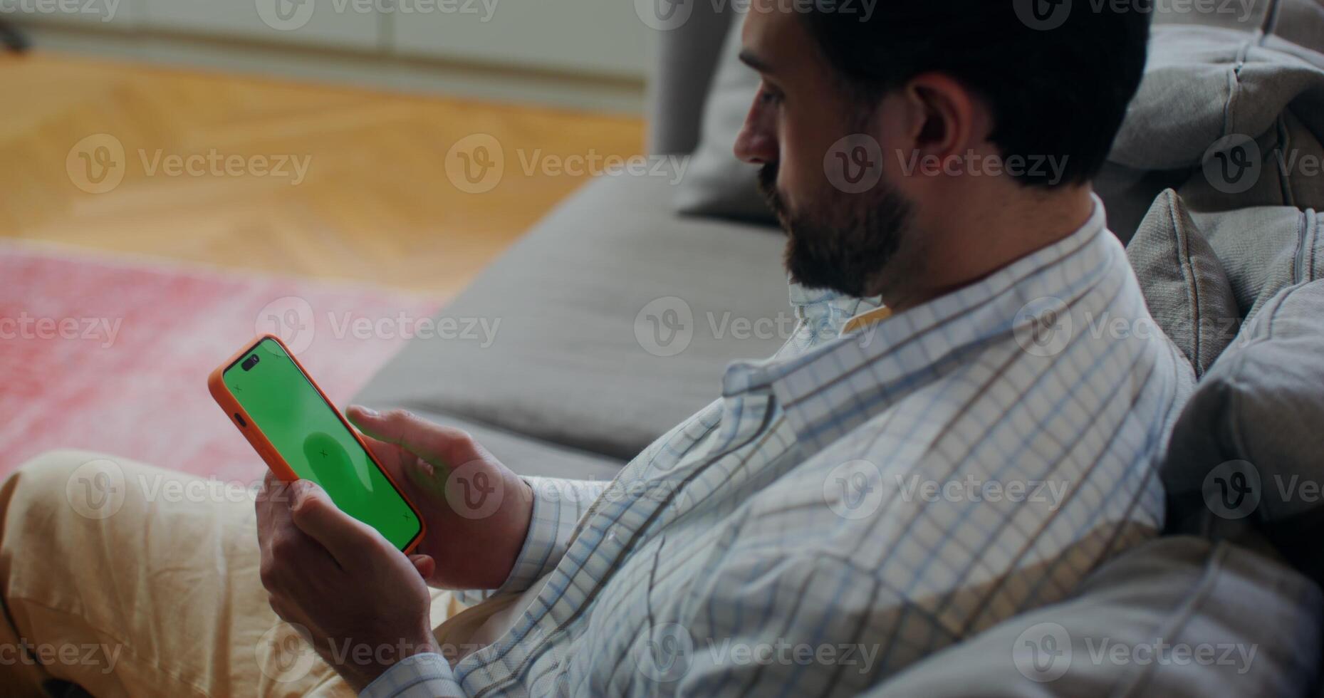 A bearded man uses a mobile phone with a green screen, scrolling the screen with his finger while sitting on a sofa indoors photo