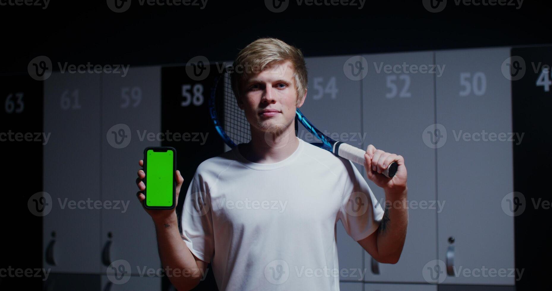 Young man of European appearance, with a tennis racket in his hands, smiling, looking straight into the camera and holding a phone with a green screen in his hands, standing in a stylish locker room photo
