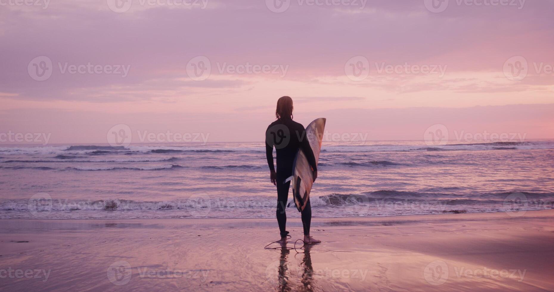 View of a man in a diving suit with wet hair and a surfboard in his hands, standing on the seashore in the pink light of the setting sun and looking into the distance photo