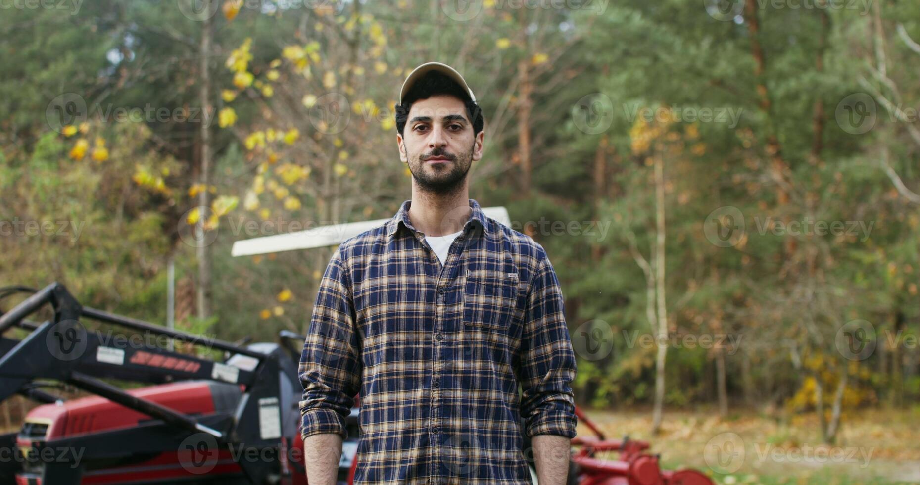 Young dressed man of European appearance in a baseball cap smiling looking straight into the camera, while standing against the background of a modern agricultural machine photo