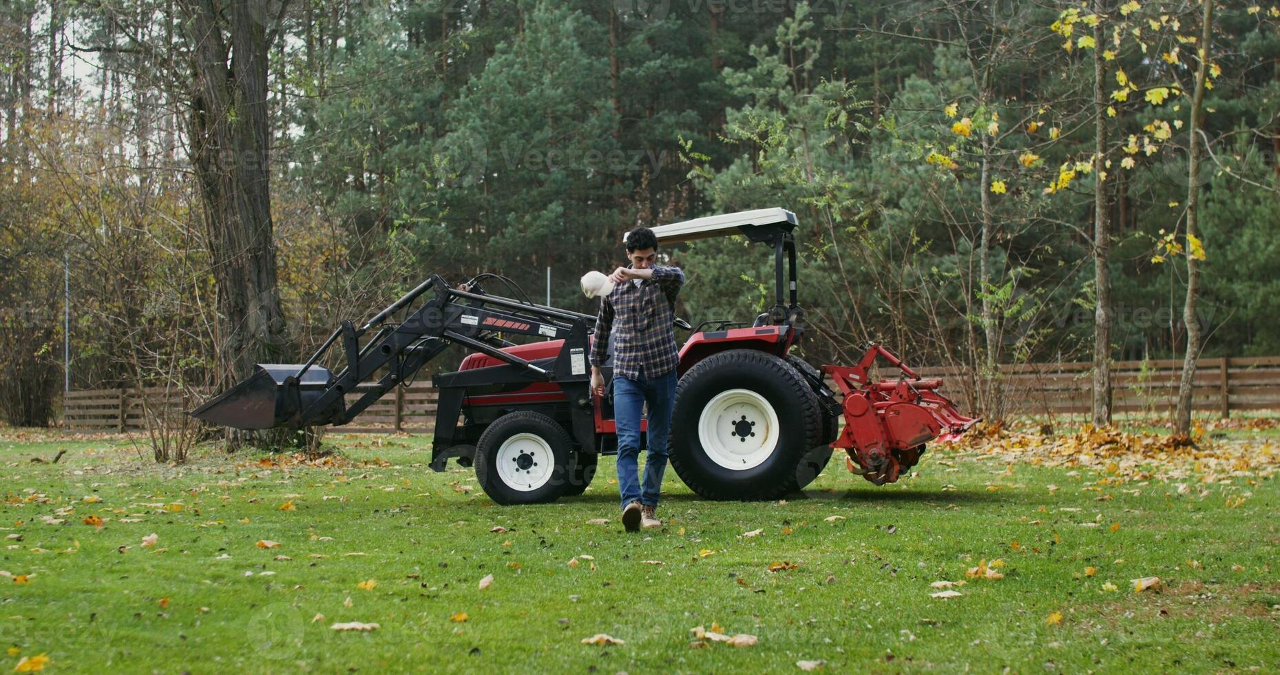 Young man climbs out of an agricultural vehicle tired after working in the field, taking off his baseball cap and wiping his forehead with his hand, on a sunny autumn day photo