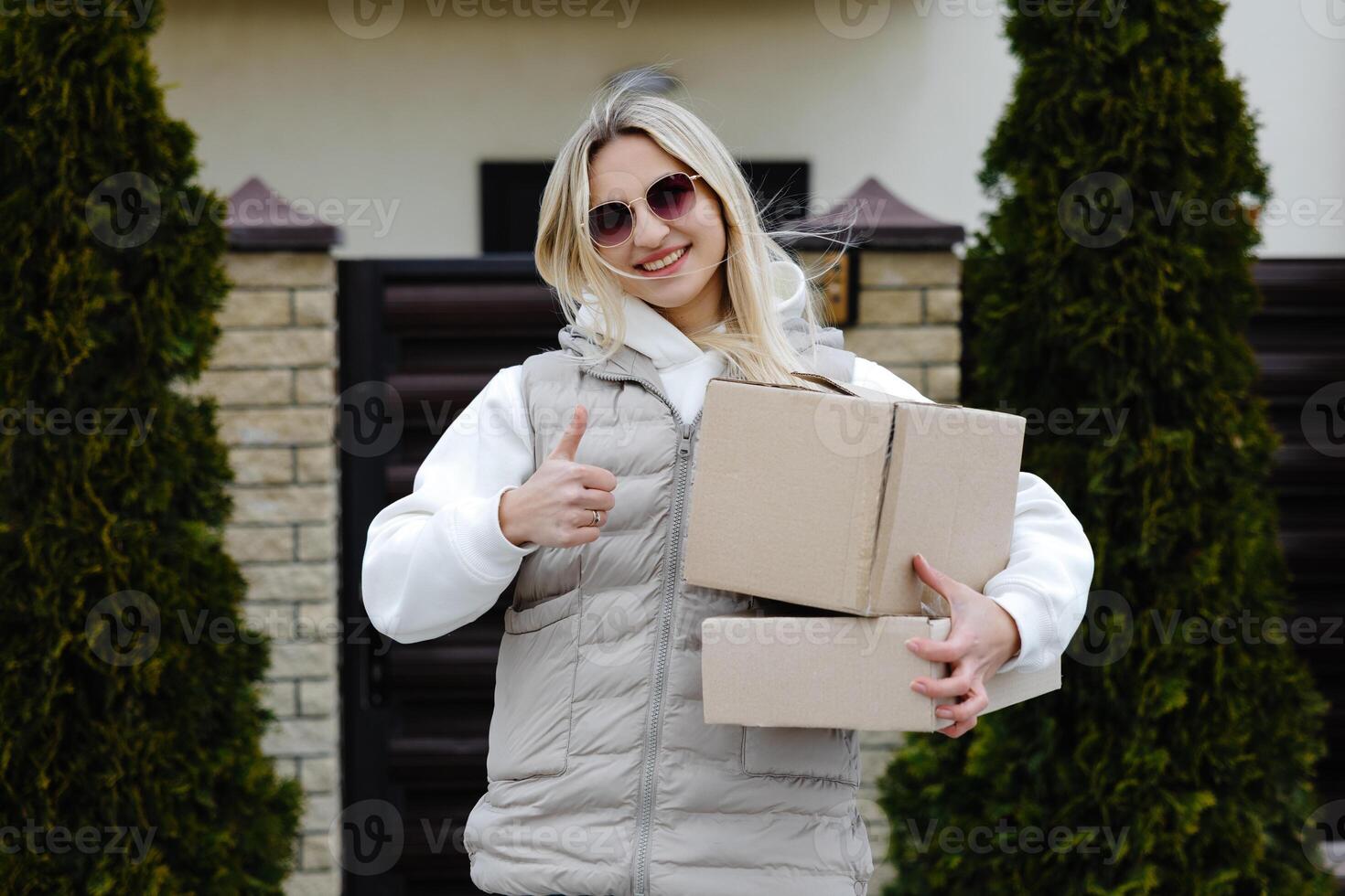 Courier showing thumbs up while holding cardboard boxes in front of a house photo