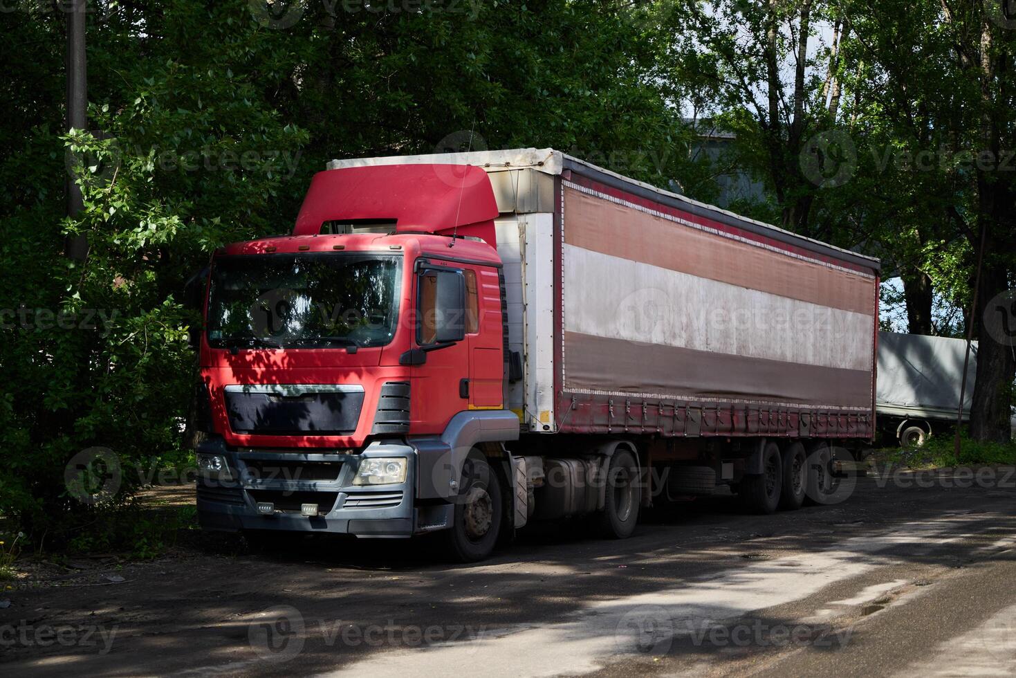 A vibrant red delivery truck is parked on the street, beautifully surrounded by lush greenery photo