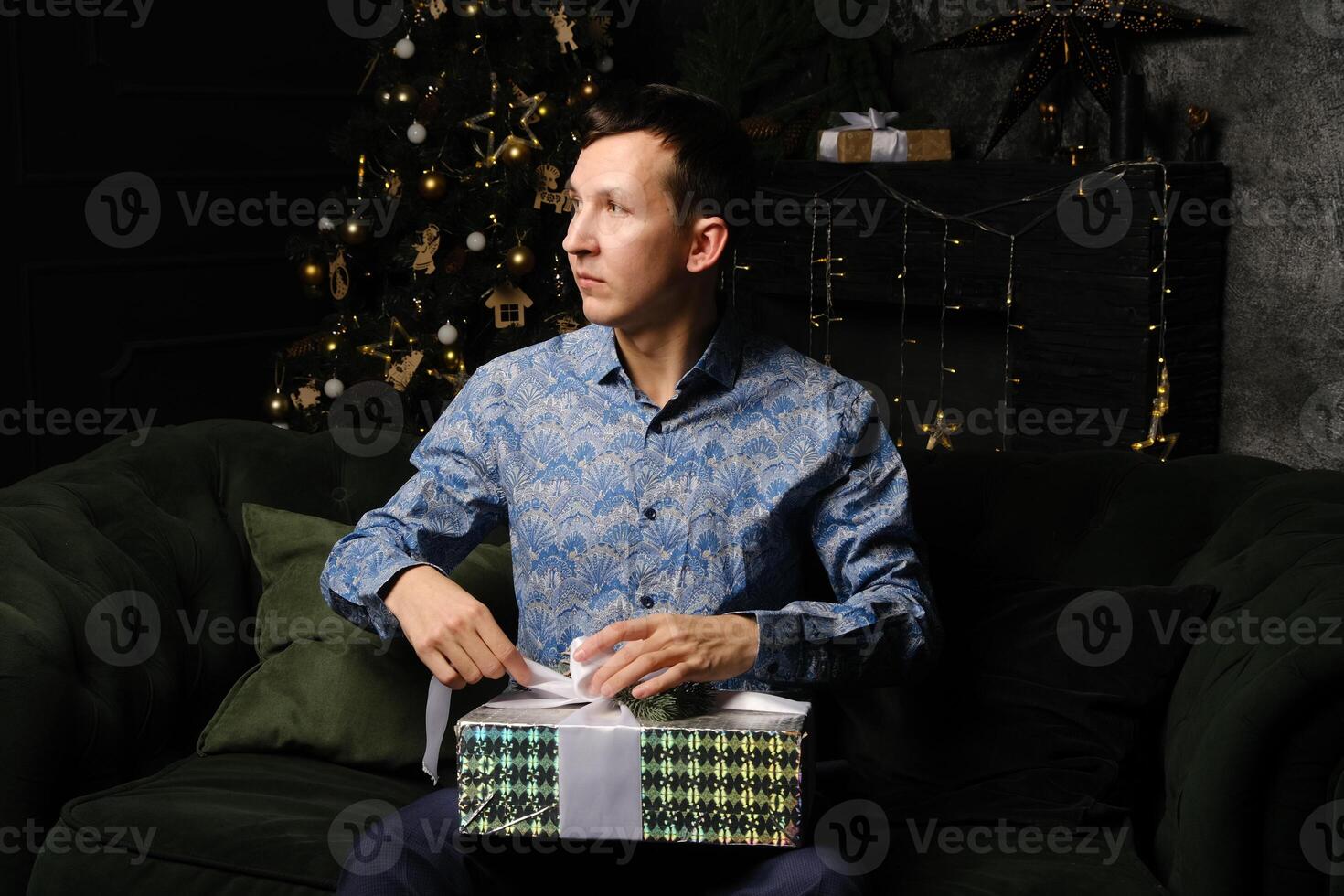 A Joyful Man Unwrapping a Gift Next to a Beautifully Decorated Christmas Tree with Bright Lights photo