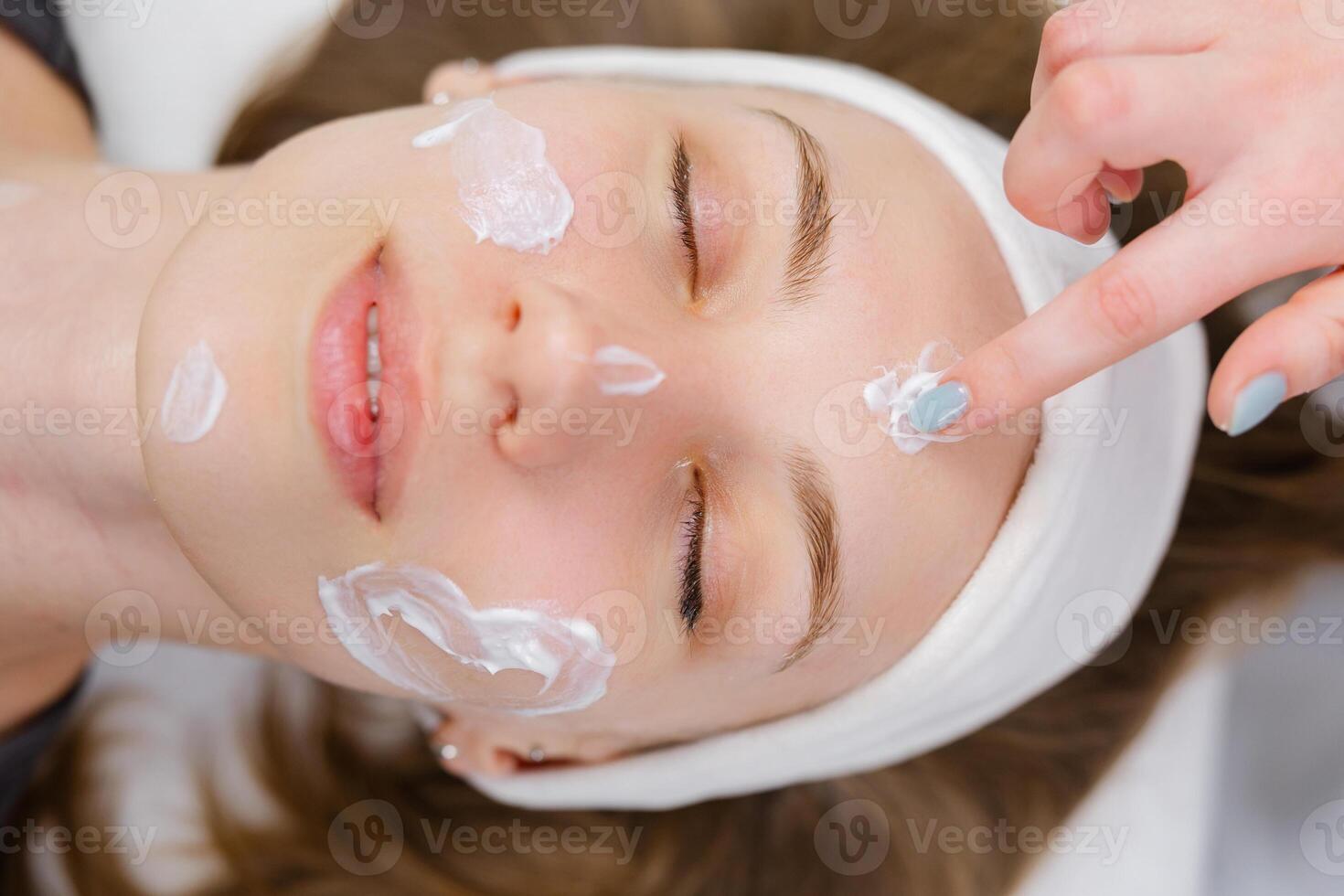 Beautician applying moisturizing cream on face of young woman lying with closed eyes photo