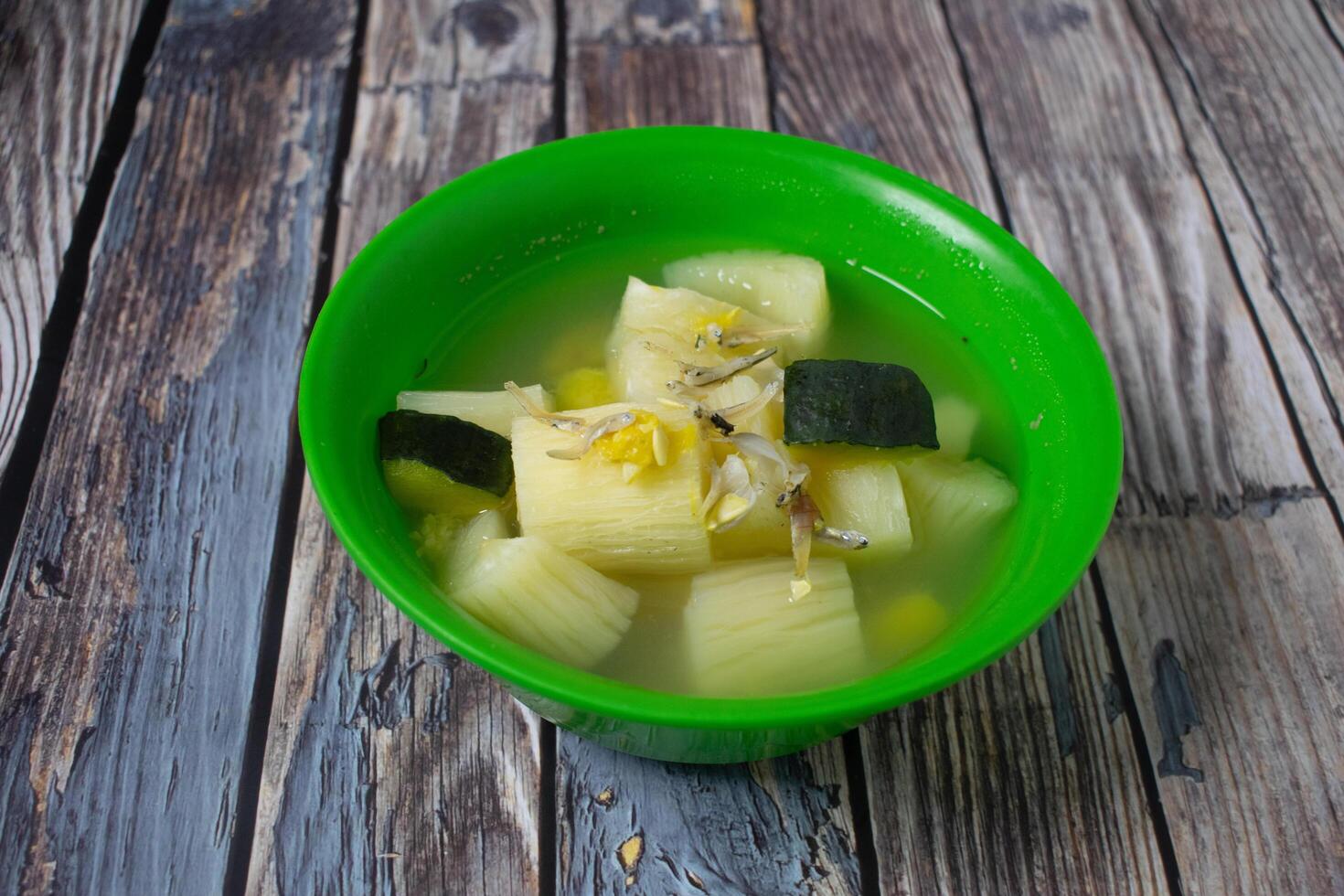 A bowl of clear vegetable soup with cassava, pumpkin and anchovies, served in a bright green bowl on a rustic wooden table. photo