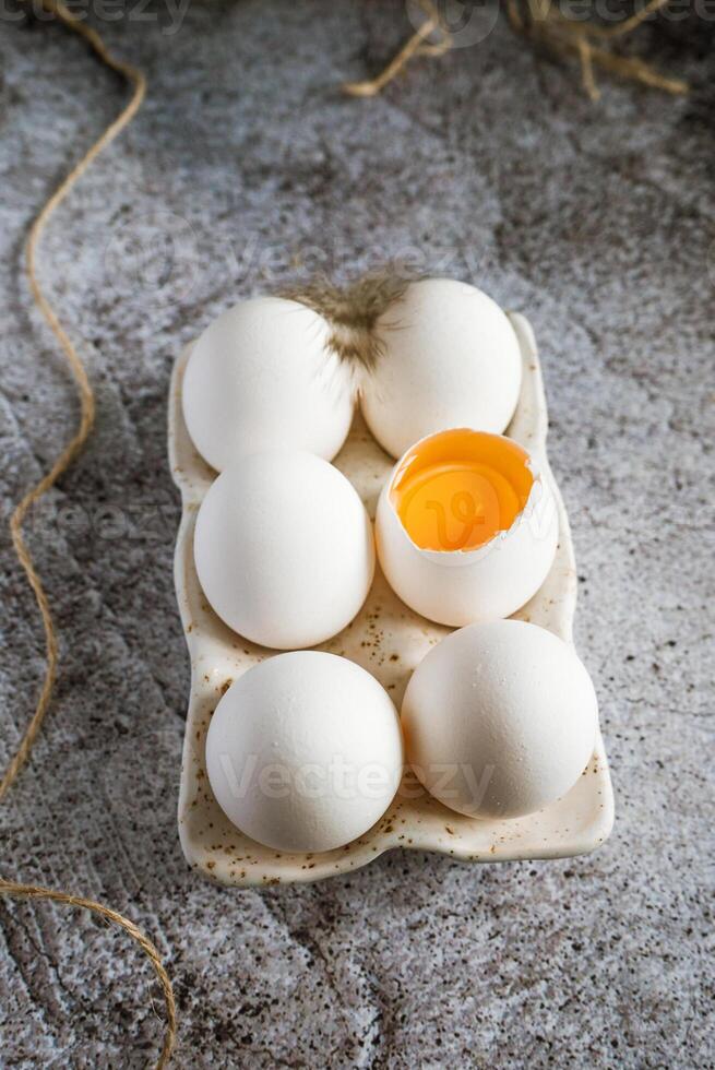 6 chicken eggs standing with feathers in a ceramic stand on a gray background, one broken egg photo