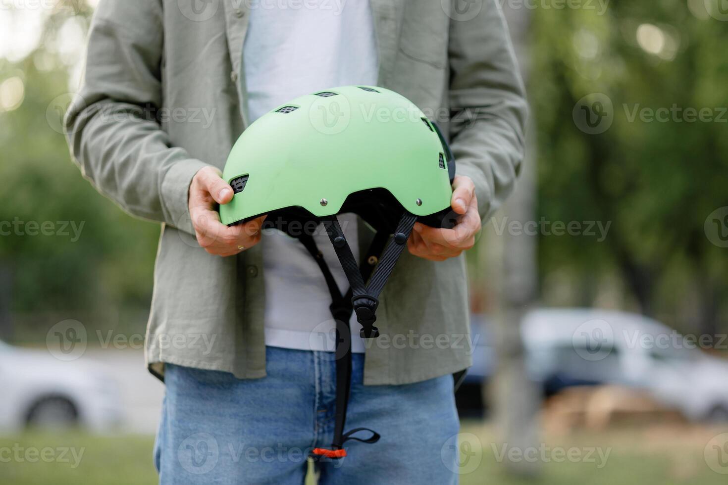 Individual holds a green helmet in a park surrounded by trees and parked vehicles during daylight hours photo