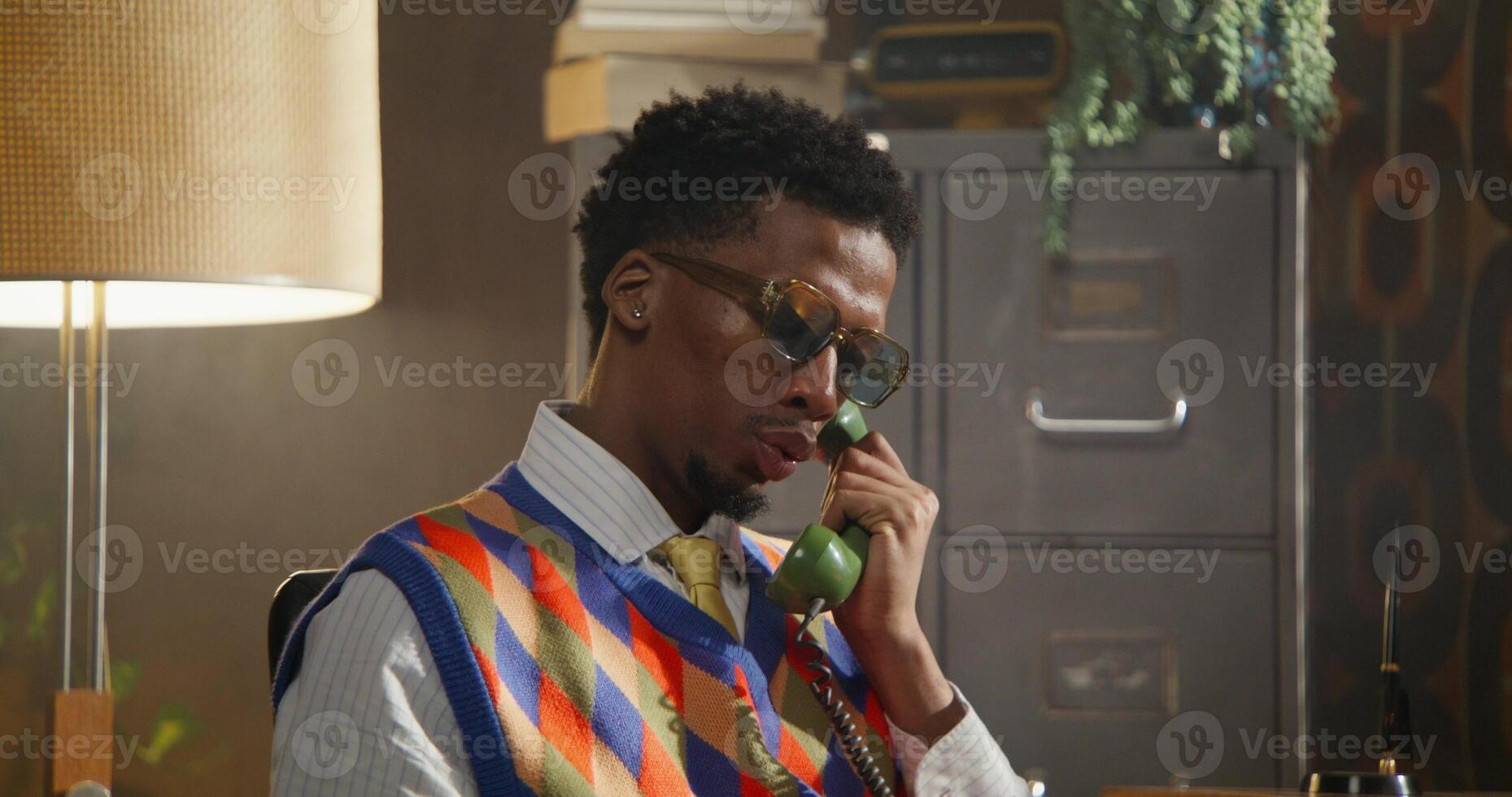 The man dials a number on a wired phone and starts talking. He's sitting in a retro office. photo