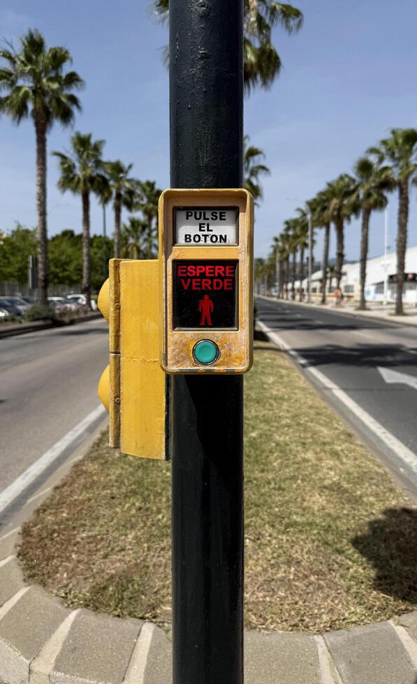 Malaga, Spain, May 18, 2025. Pedestrian traffic signal button in yellow casing instructs to press for crossing, set against a street lined with palm trees and clear blue sky, indicating urban life photo