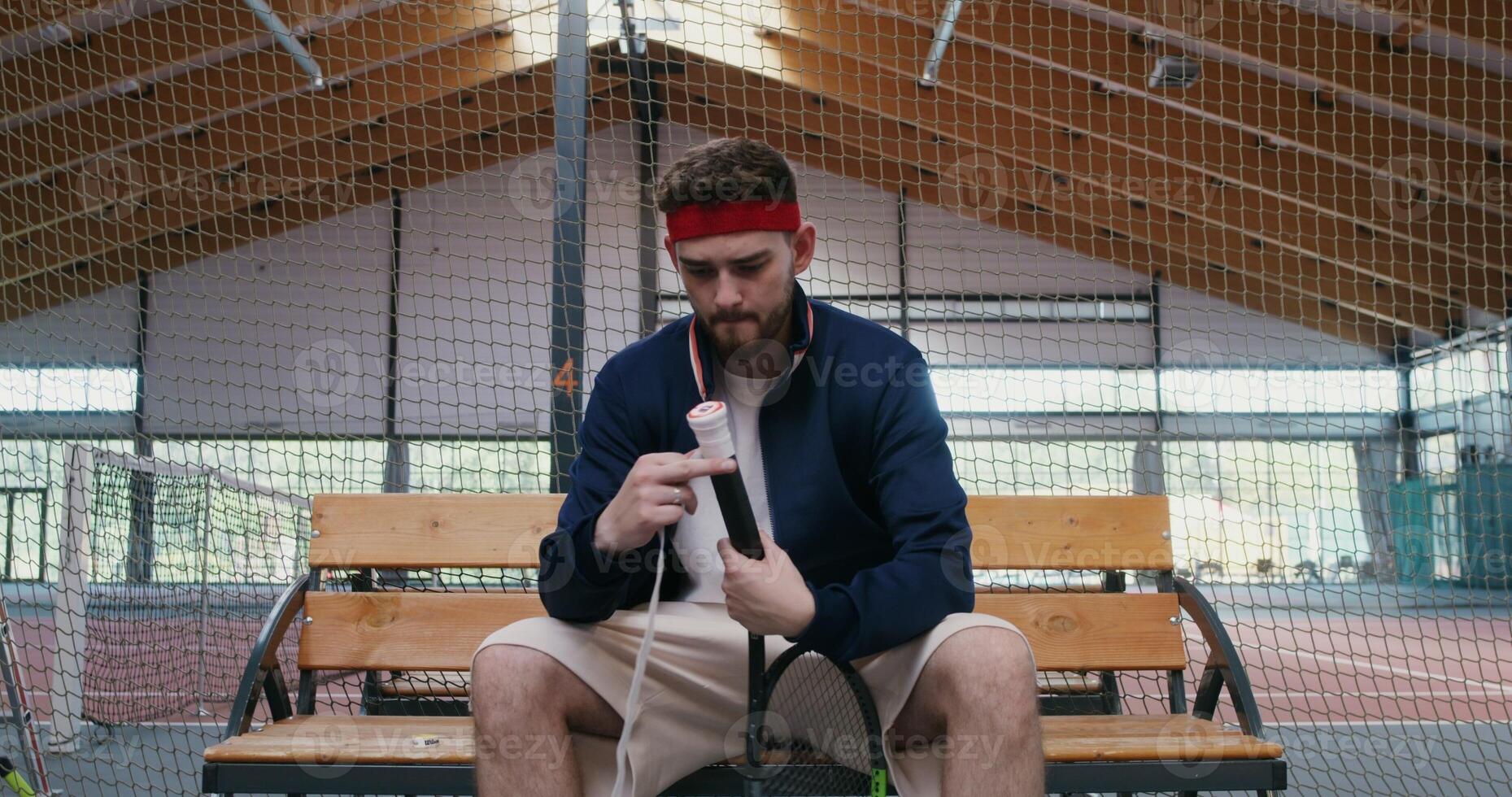 Young man of European appearance wraps the handle of a tennis racket with a special tape for better grip on the hand, sitting on a bench near the tennis court, preparing for a tennis match, close-up photo