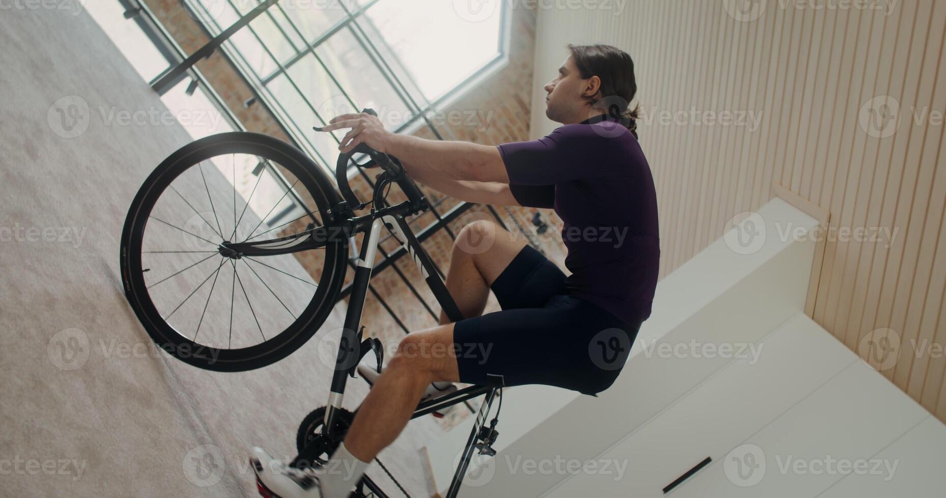 Young focused man rides a bike simulator in modern home interior. Vertical filming transforms into horizontal, circular camera movement photo