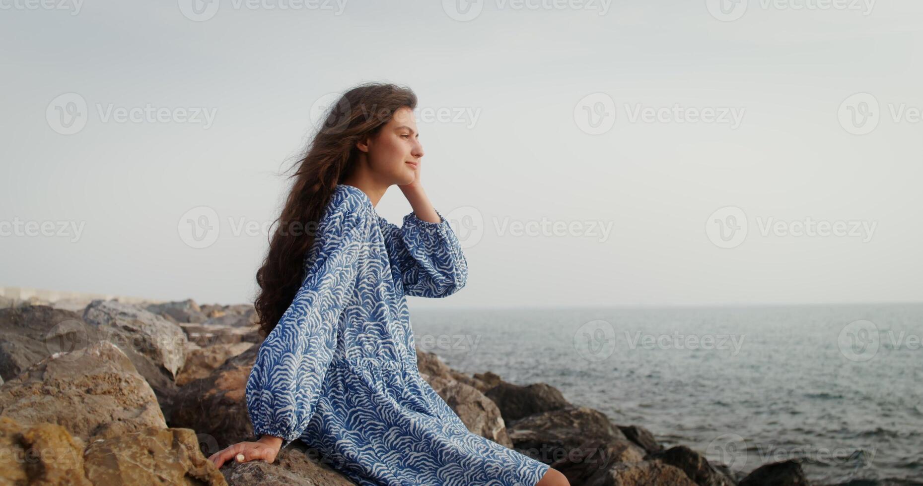 Woman straightens her thick dark hair, looking with pleasure into the sea distance, sitting on the stones on the seashore. 4k , red komodo photo
