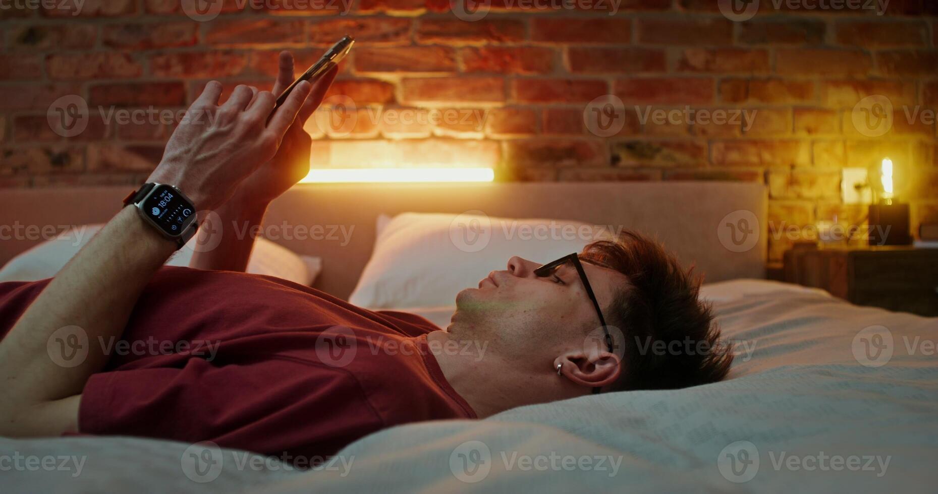 A young man in glasses dressed in casual clothes uses smartphone while lying across the bed in a stylish bedroom in the evening photo