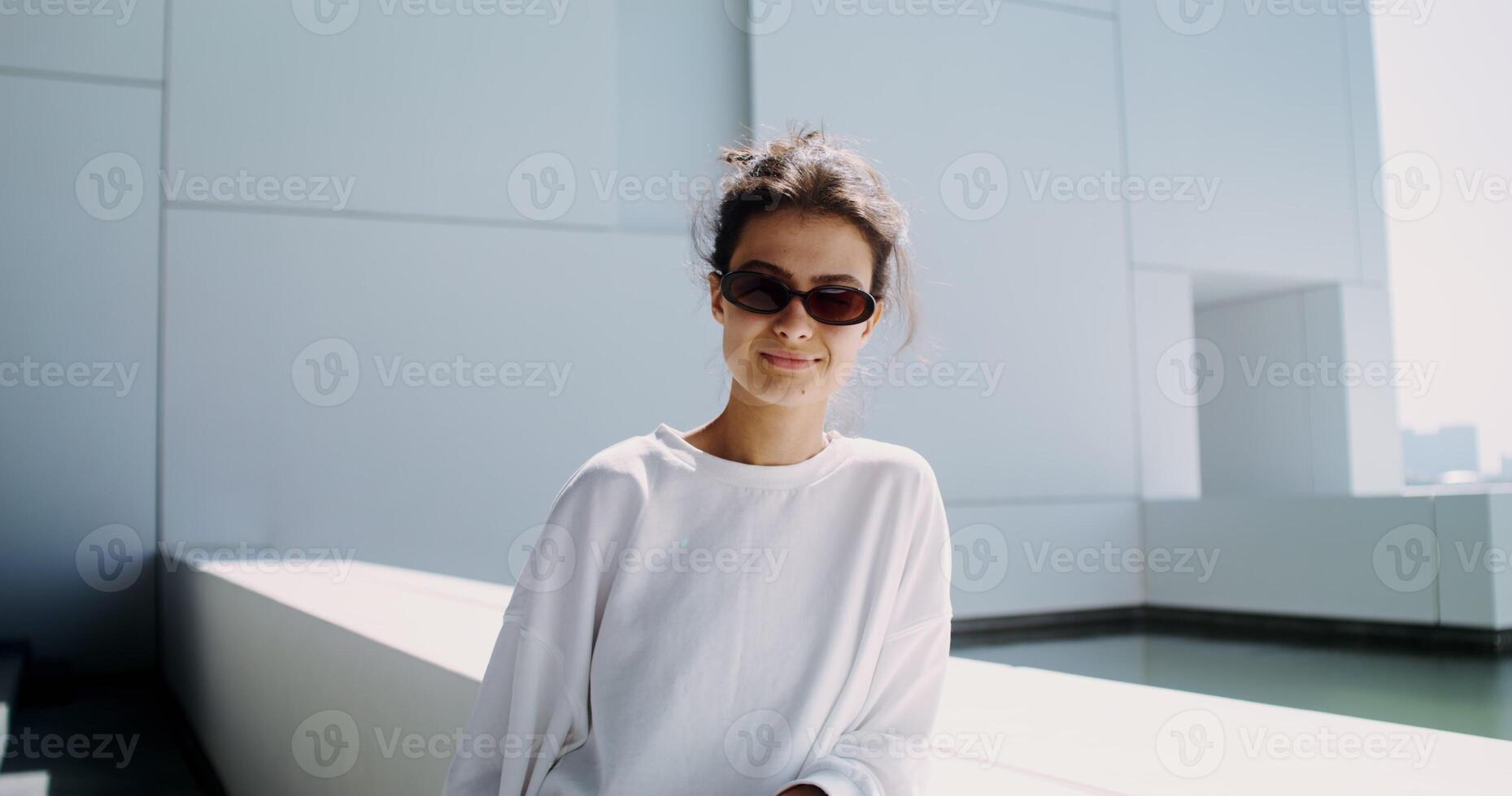 A young woman in sunglasses, with her hair gathered, stands on the balcony of a modern minimalist white building photo