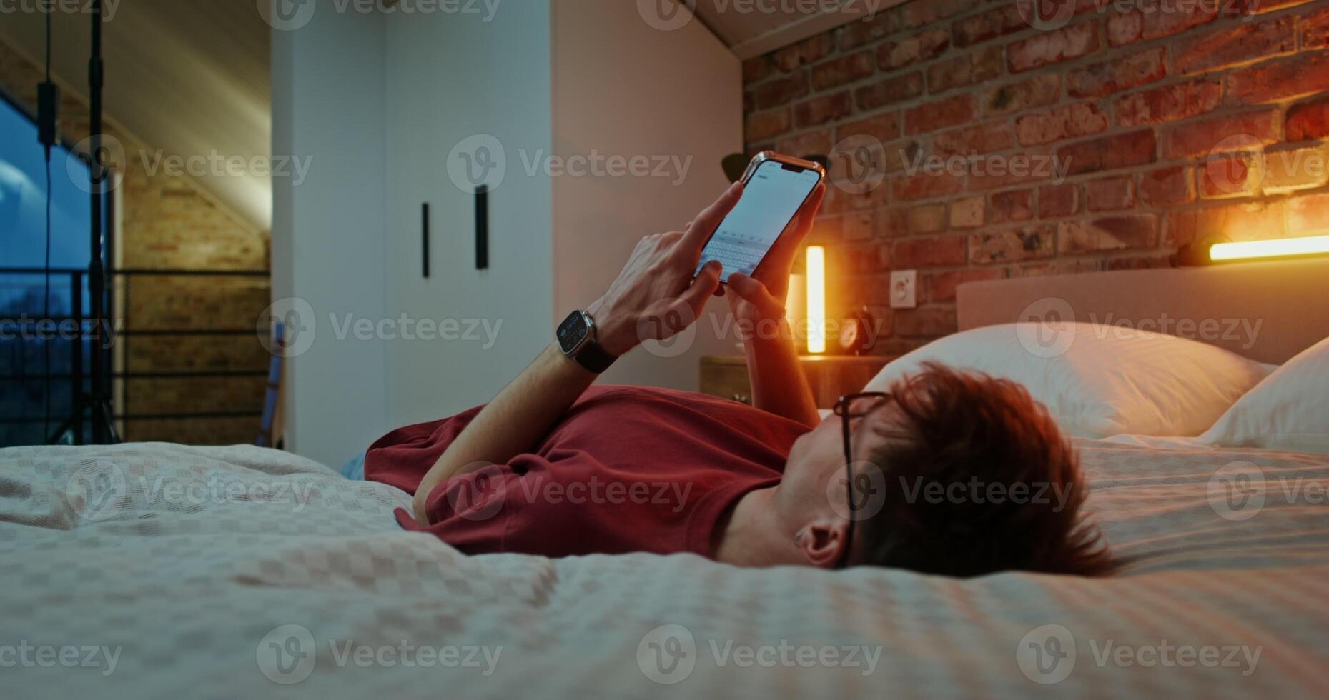Young man in glasses dressed in casual clothes is typing text on a mobile phone, while lying across the bed in the evening in a stylish home interior photo
