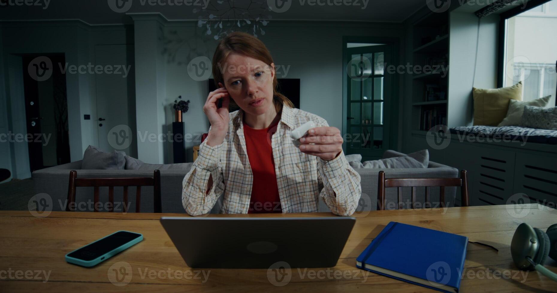 Red-haired woman puts on headphones while talking on a chat on a laptop, sitting at a table in a stylish home interior with a serious look, working remotely from home photo