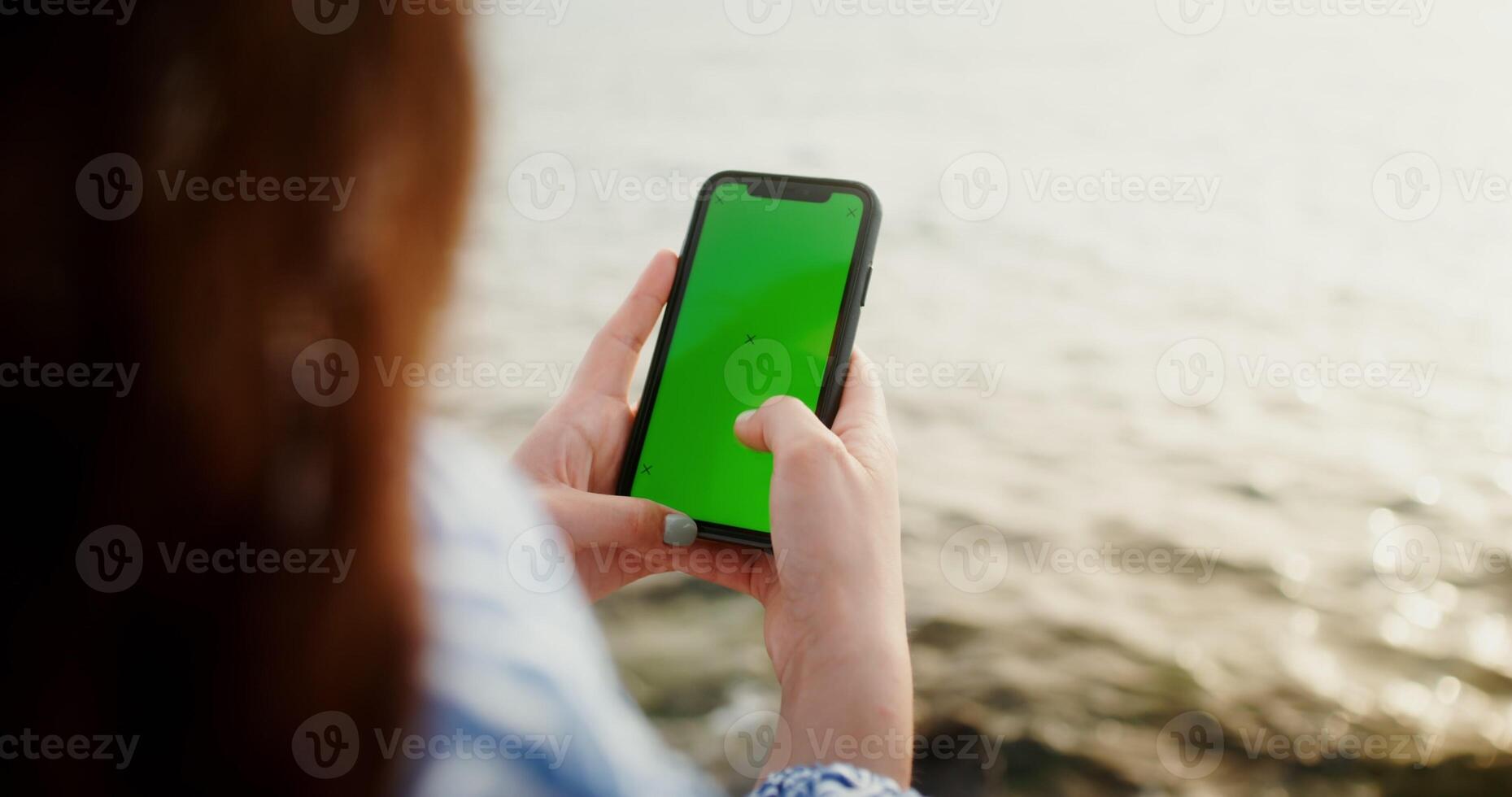 Close-up of phone with a green screen in the hands of a woman typing on a smartphone while sitting on the rocks on the seashore. Chroma key, mockup, copy space. 4k , red komodo photo