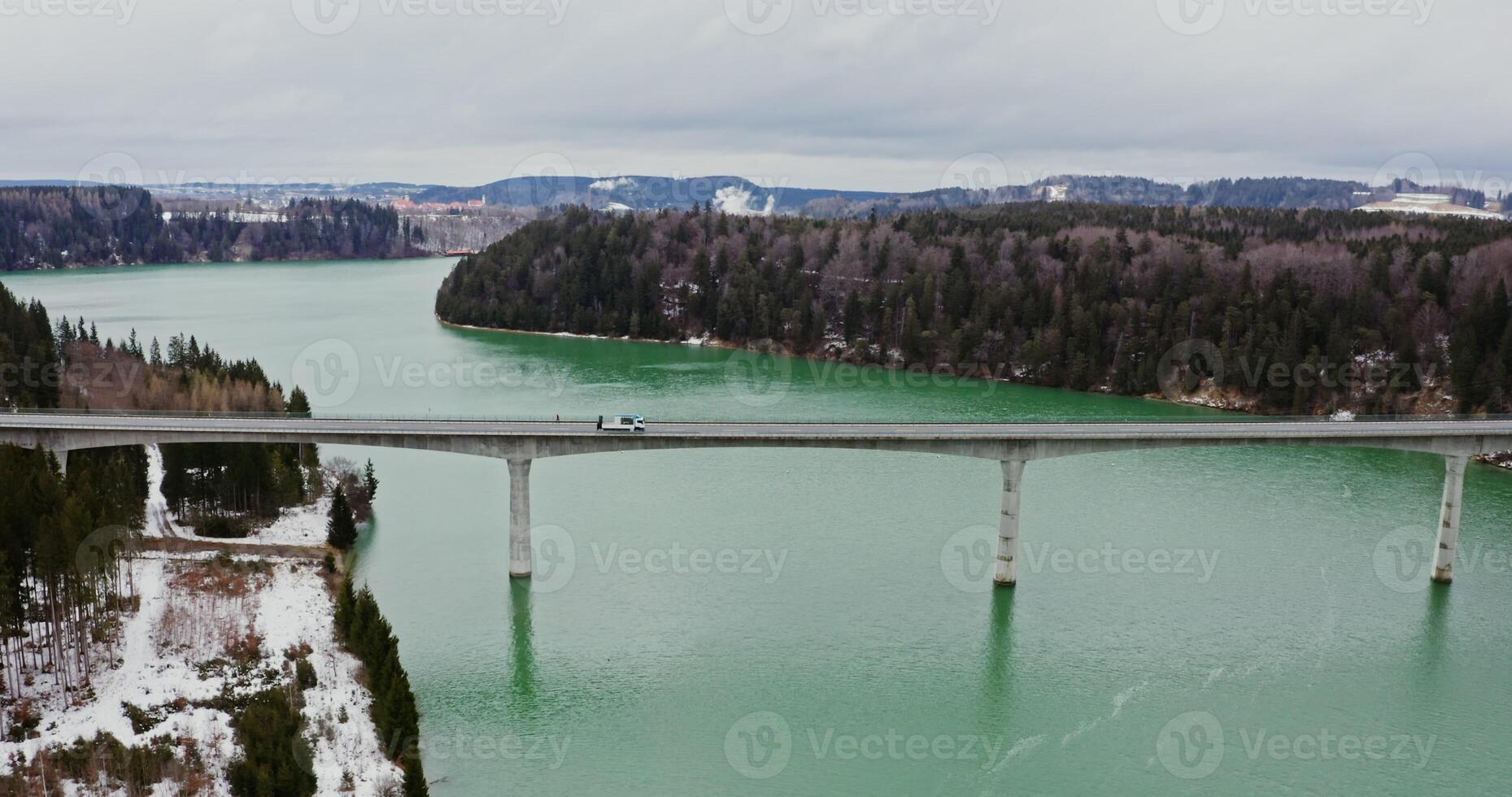 Concrete bridge for road transport across a wide river in a picturesque place among forests and fields covered with snow, drone , from the side of the river photo