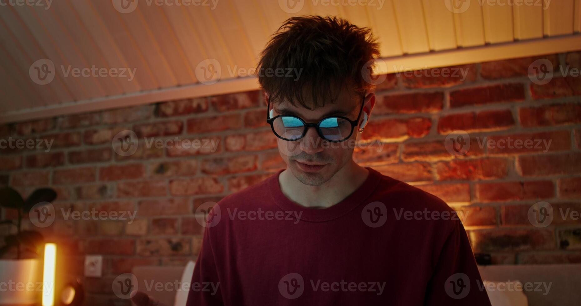 Young stylish man with headphones in his ears talking on chat on laptop, while sitting in lotus position on bed in the evening photo