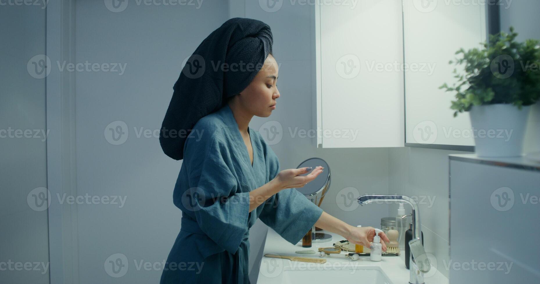 Beautiful woman of Asian appearance in a bathrobe, with a towel on her head, applies a cosmetic product on her face while looking in a mirror in a stylish bright bathroom photo
