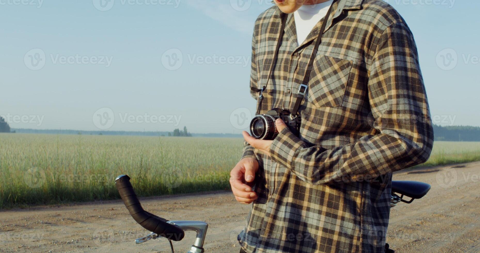 A young stylishly dressed man takes photographs with a camera while standing near a picturesque wheat field on a sunny day photo