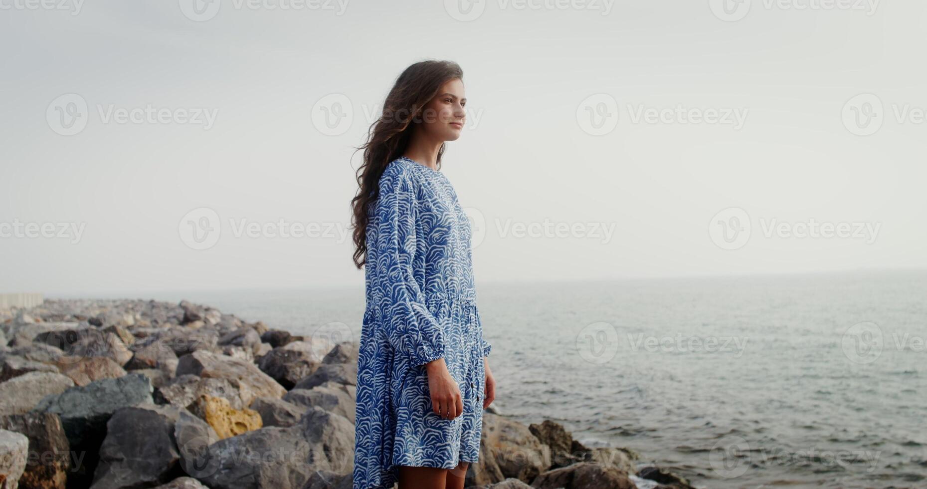 A beautiful woman of European appearance with curly black hair stands on large stones on the seashore and smiles looking into the distance. 4k , red komodo photo
