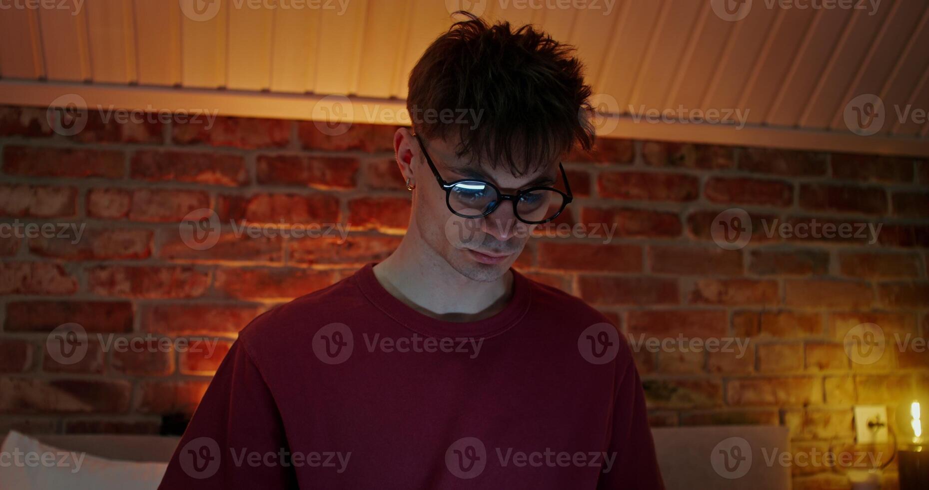 A young stylish man looks down at the screen of a laptop or tablet while sitting in the bedroom on the bed. Close-up of a concentrated man's face photo