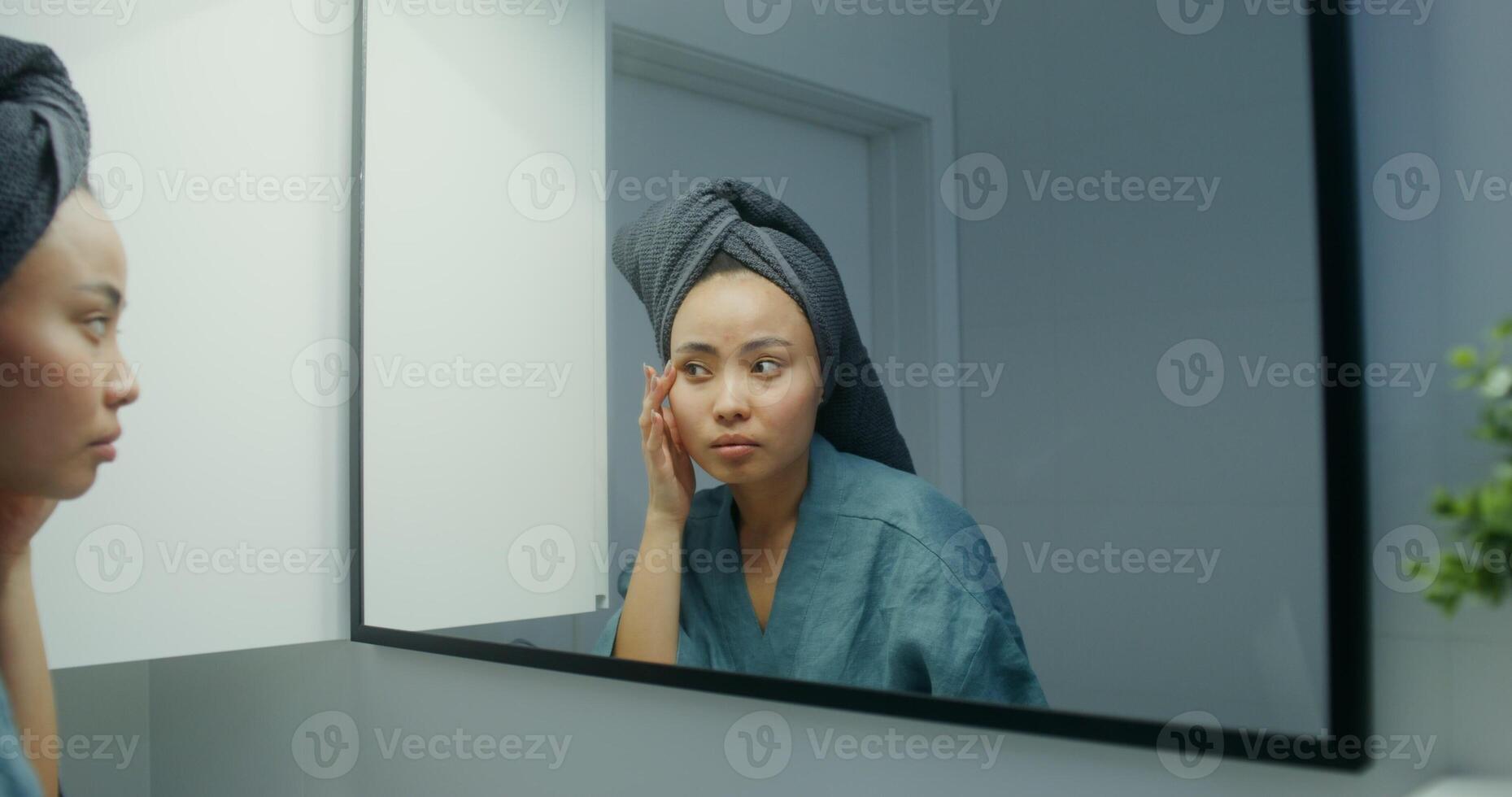 Young Asian woman in bathrobe, with a towel on her head, touches her face while looking in the mirror in a bright bathroom. The moves from the woman's face to the general shot and back photo