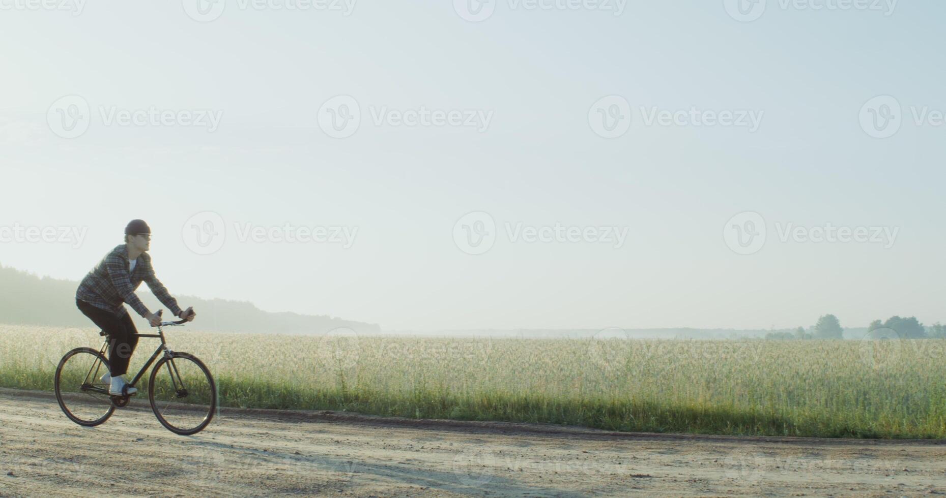 A young stylishly dressed man rides a bicycle on a gravel road near a wheat field in the rays of the rising sun. 4k , red komodo photo