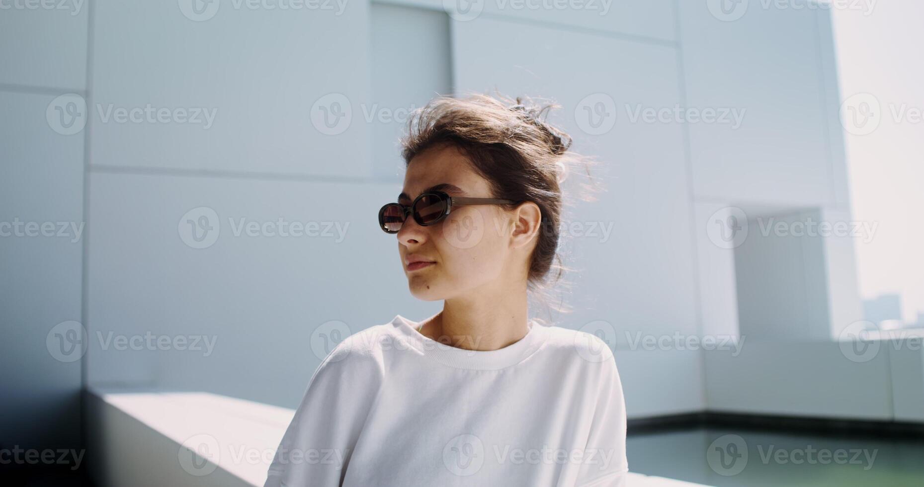 A woman in casual clothes, wearing sunglasses, with her hair gathered, stands on the balcony of a modern minimalist white building surrounded by water photo