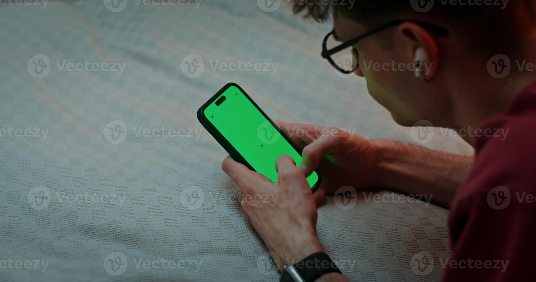 Man is typing on a smartphone with a green screen while lying on his stomach on the bed, top view. Chroma key, copy space photo