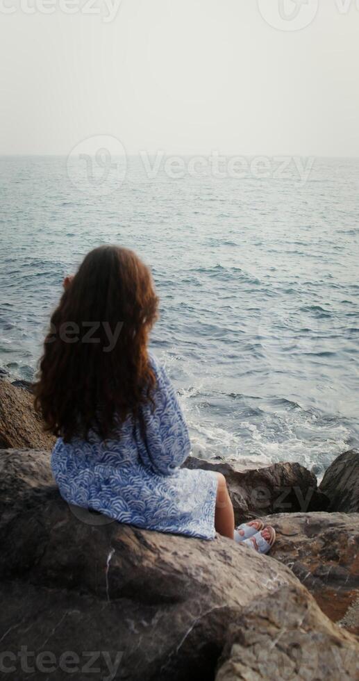 Woman straightens her wavy dark hair, sitting on the rocks on the seashore on a sunny day and looking into distance. Vertical from back, no face, unrecognizable person. 4k , red komodo photo