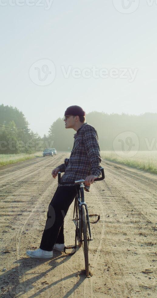 A young man with a camera around his neck stops on a gravel road near field after cycling and admires the beauty of the surrounding fields. 4k vertical , red komodo photo