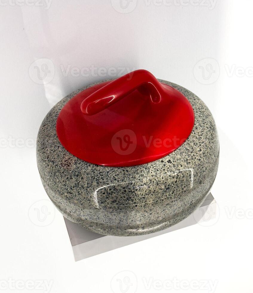 This high-angle, close-up shot captures a traditional curling stone with a vibrant red handle, set against a pristine white background. The granite body of the stone shows its unique speckled texture photo