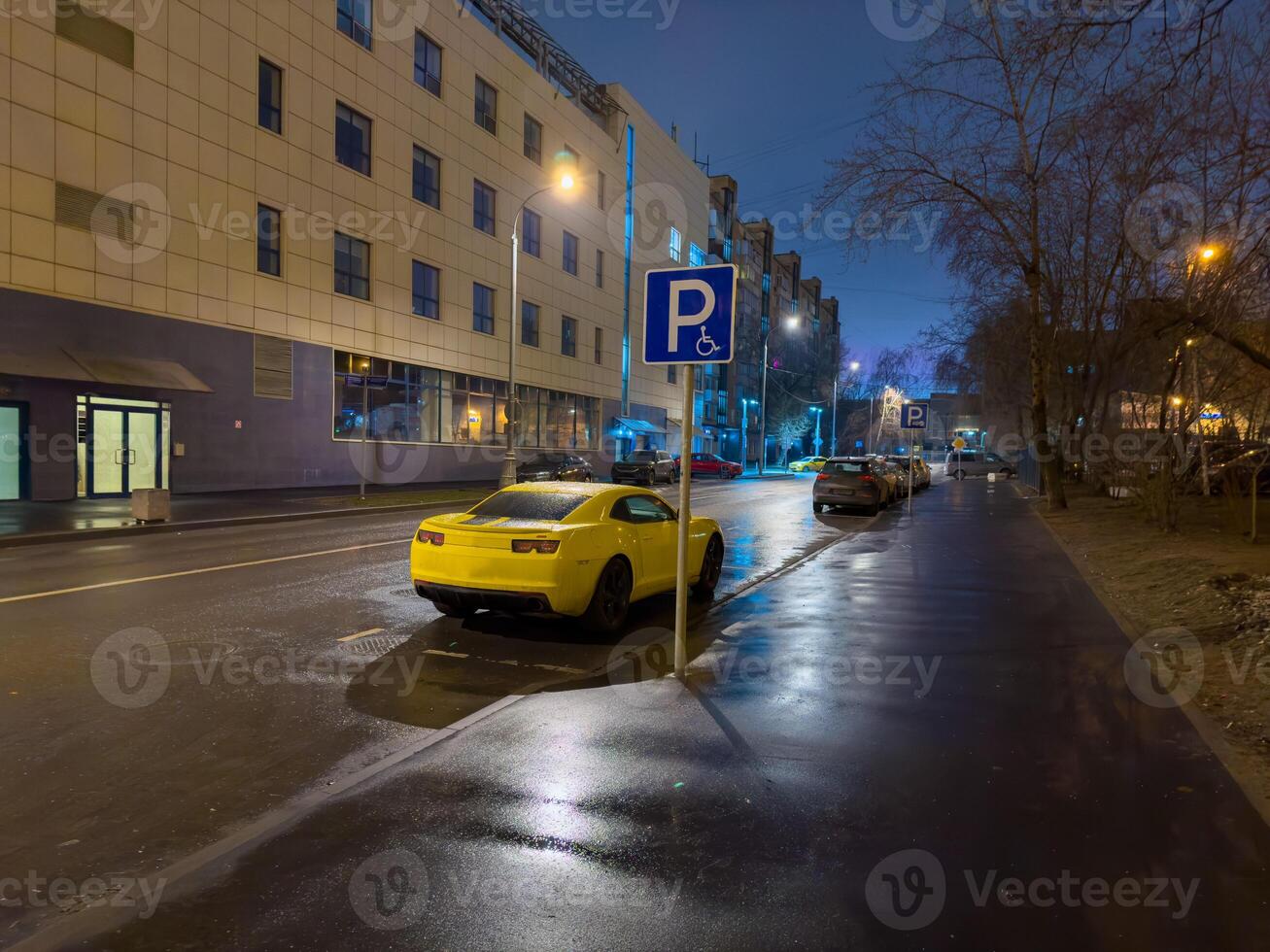 A vibrant yellow sport car is conspicuously parked on a wet city street at dusk, reflecting the glowing streetlights and the illuminated facade of a modern building. The low-angle shot captures the photo