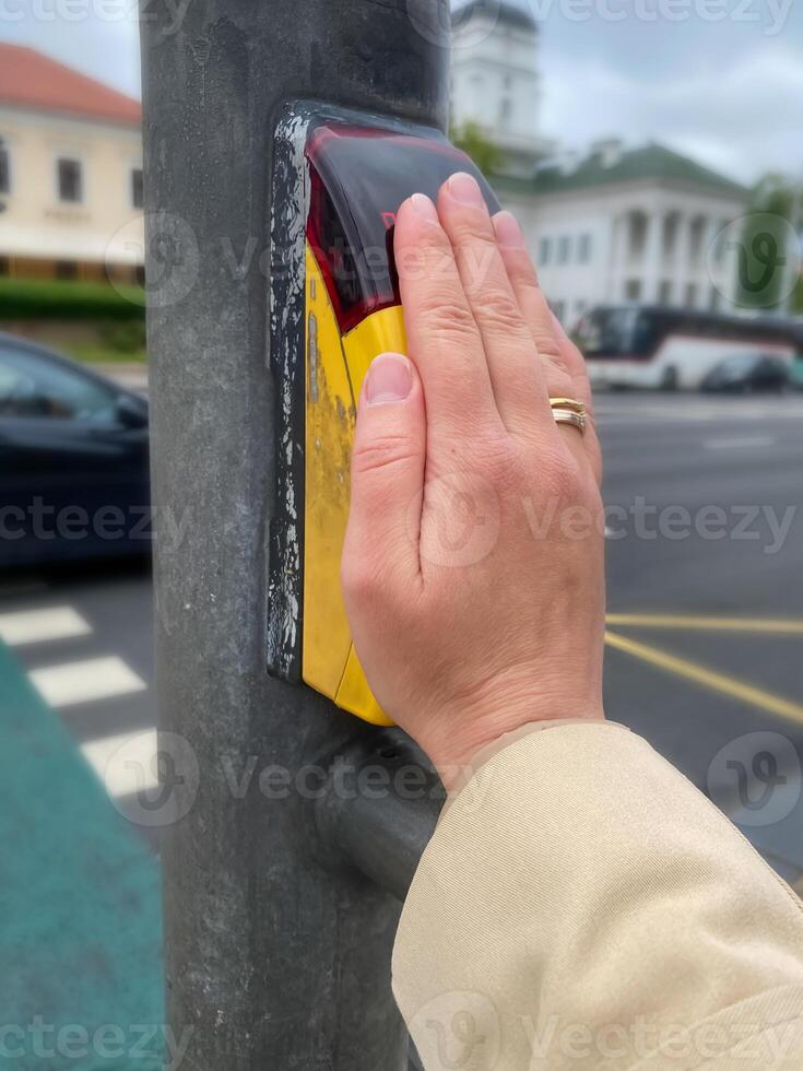 Female hand pressing pedestrian crossing button on a traffic light pole, urban street setting photo