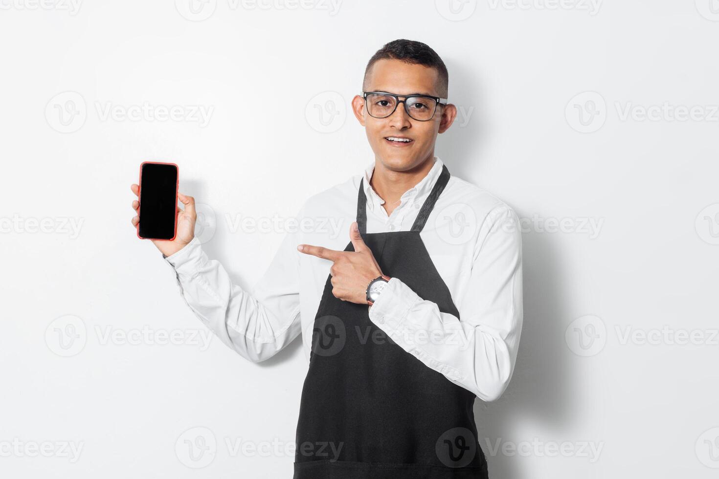 Waiter young hispanic man with apron holding and pointing with a finger a mobile. photo