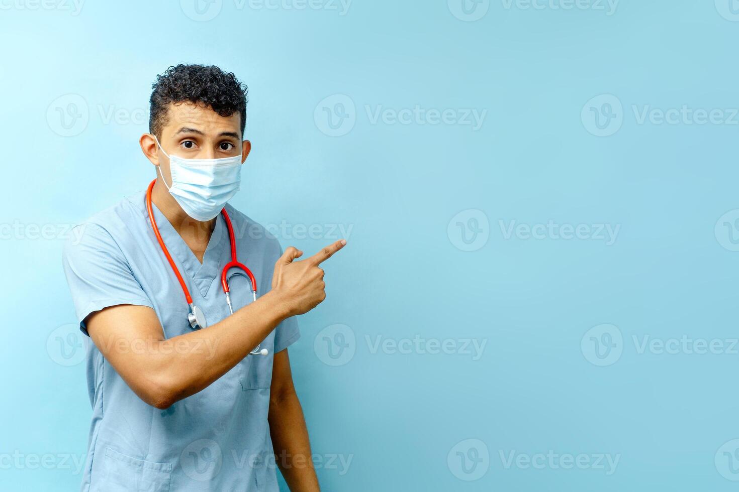 Hispanic man with face mask with hand points to something on a blue background. photo