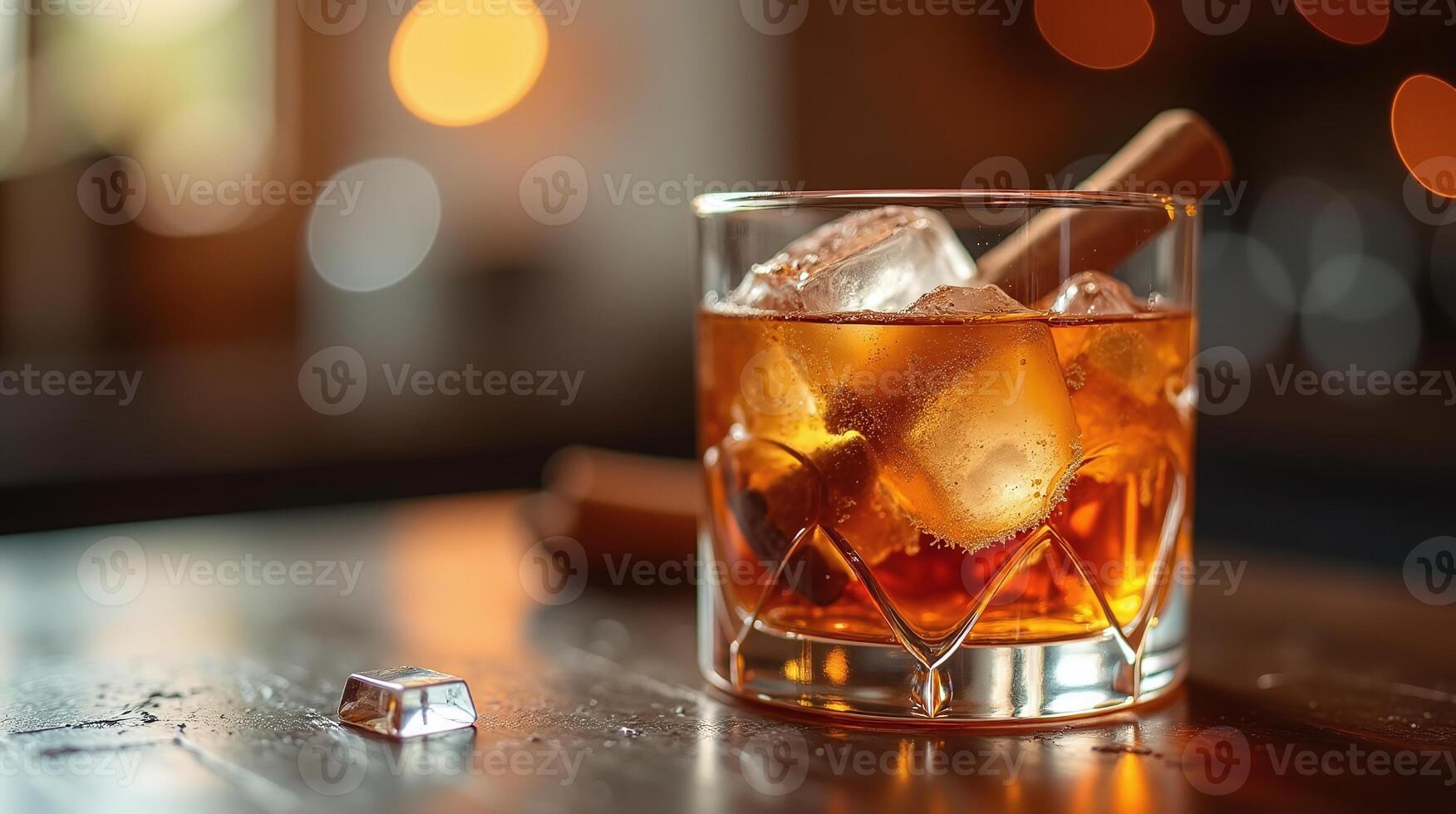 A glass of amber-colored beverage with ice, a cinnamon stick, and a cube on the table. photo