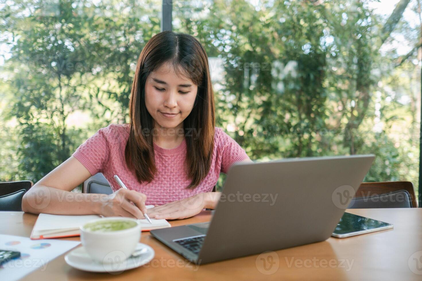 A young woman in a pink shirt works on her laptop and takes notes in a cozy cafe surrounded by lush greenery. photo