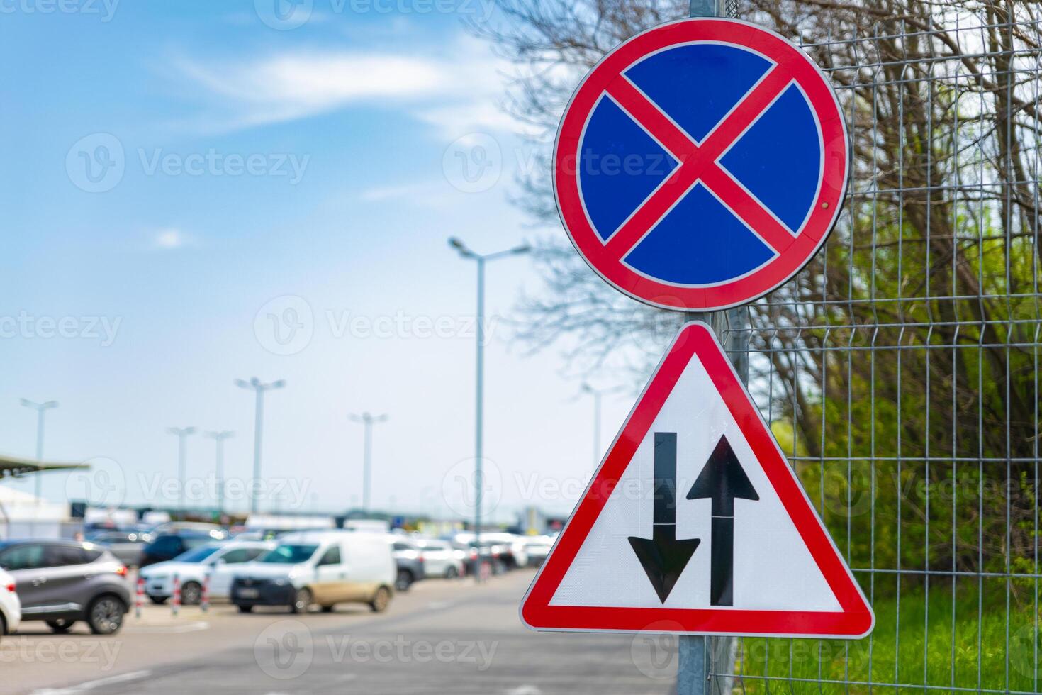 Roadside signs indicating no parking and two-way traffic, mounted on a fence near a busy parking area under a clear sky. photo