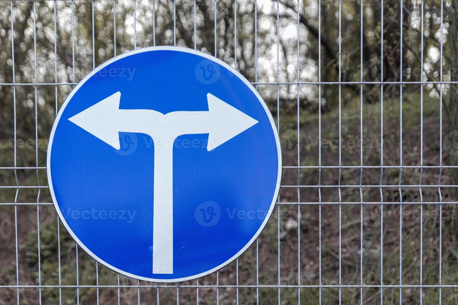 Blue circular road sign indicating a mandatory left or right turn, mounted on a wire fence with a wooded area in the background. photo