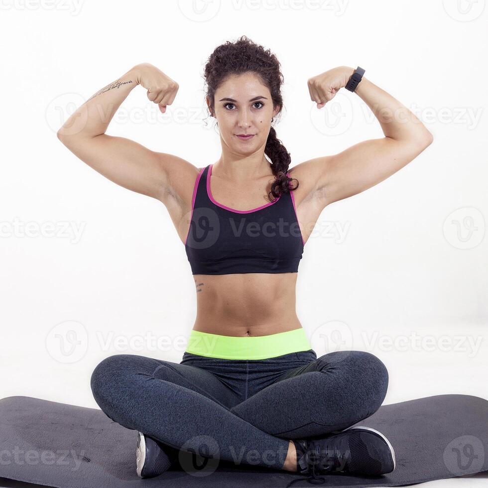 beautiful young sportswoman sitting on mat with crossed legs showing biceps isolated on white background photo