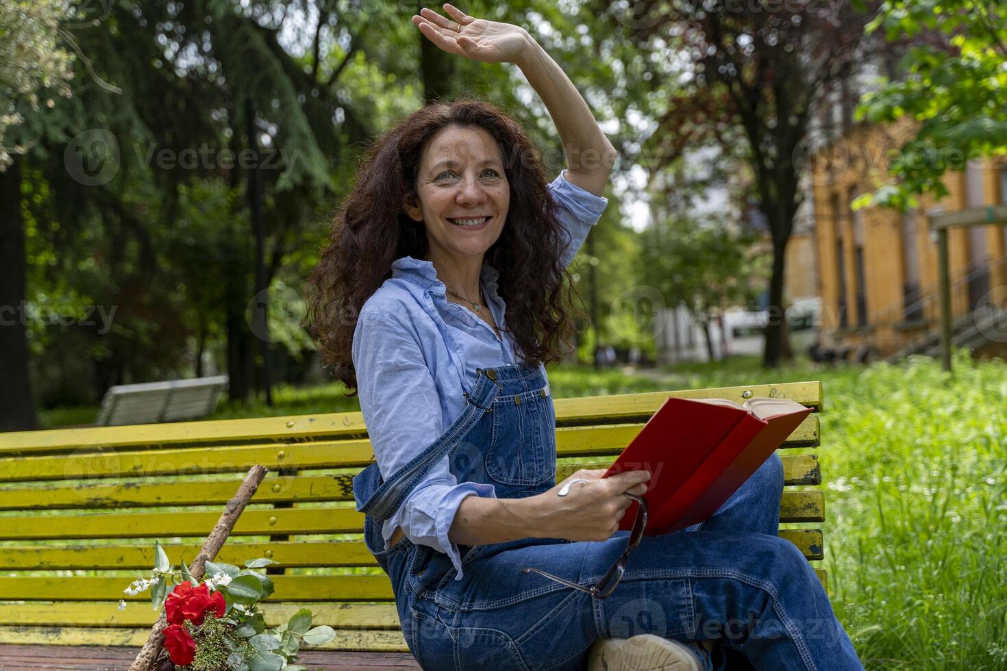 middle aged woman sitting on a yellow bench waving while reading a book photo