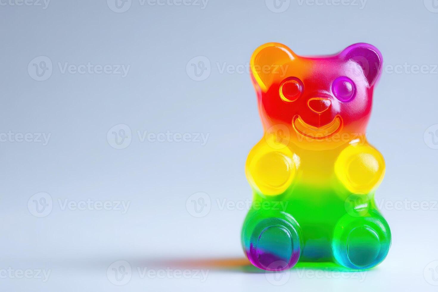 A bright gummy bear candy with a rainbow gradient sits upright on a clean, smooth surface. Its cheerful expression and vivid colors create a playful and appealing look, perfect for sweet treats. photo