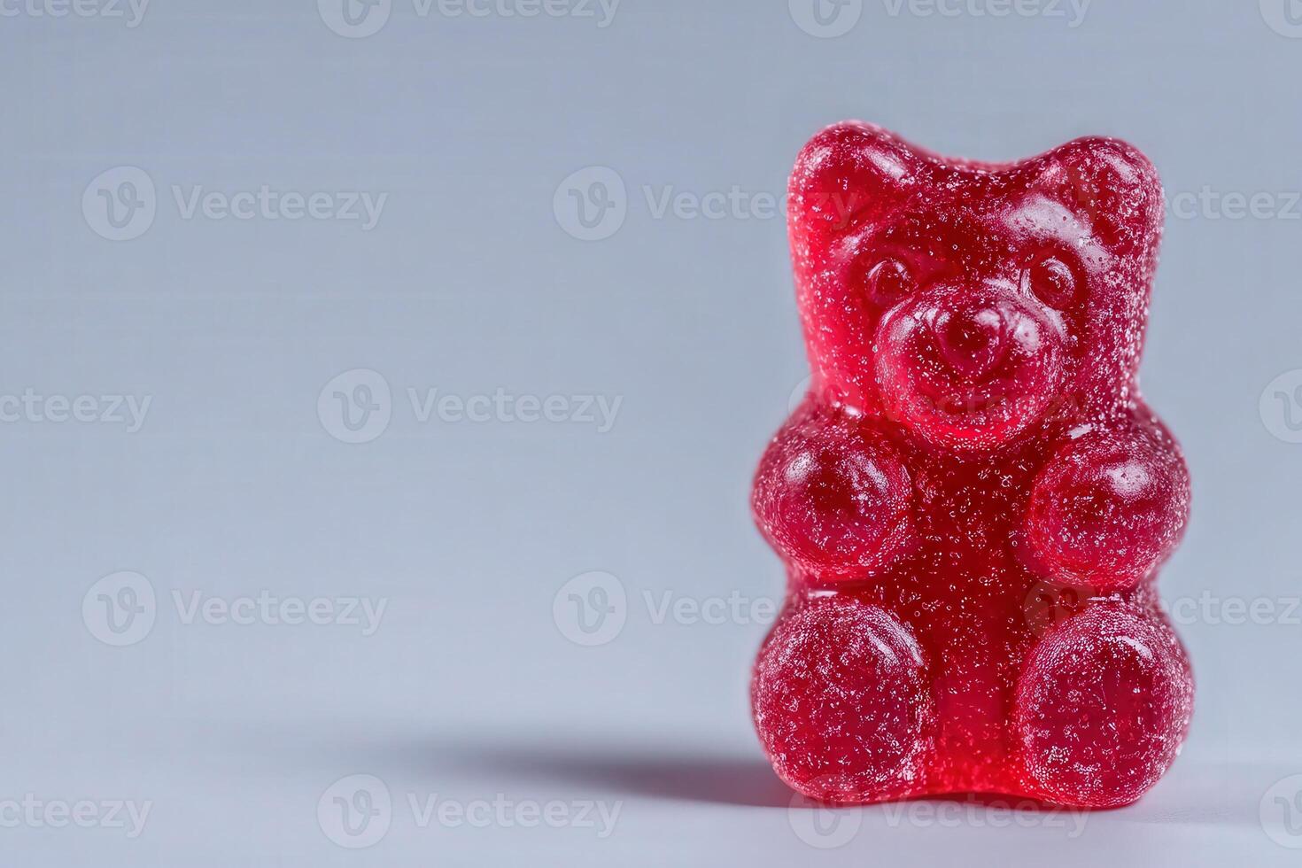 A bright red gummy bear figure, plump and textured with sugar. It sits upright on a flat surface, highlighting its cheerful expression. The neutral background enhances its vibrant color. photo