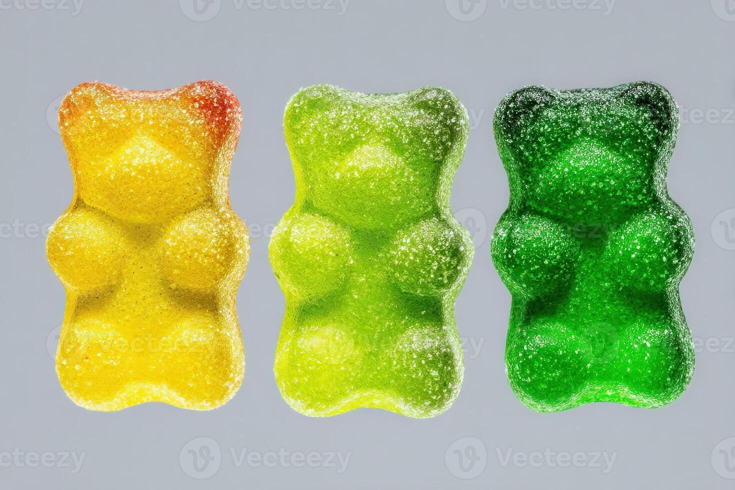 Three gummy bears in bright colors yellow, green, and a lighter green are lined up against a neutral grey background. Their sugary coating glistens softly under the light. photo