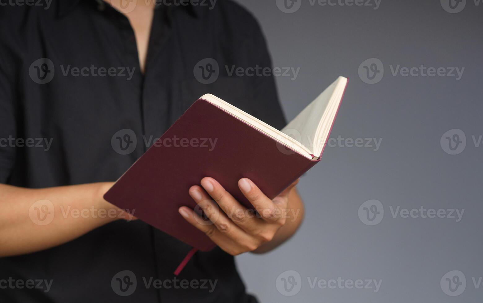 A man in a black short-sleeve shirt reads a book while standing on a gray background photo
